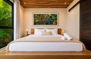 Een groot bed met een witte spreidek en kussens, een schilderij van vogels aan de muur, en een raam met gordijnen.