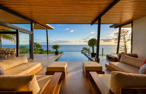 A modern infinity pool with a view of the ocean, surrounded by wooden furniture and potted plants.