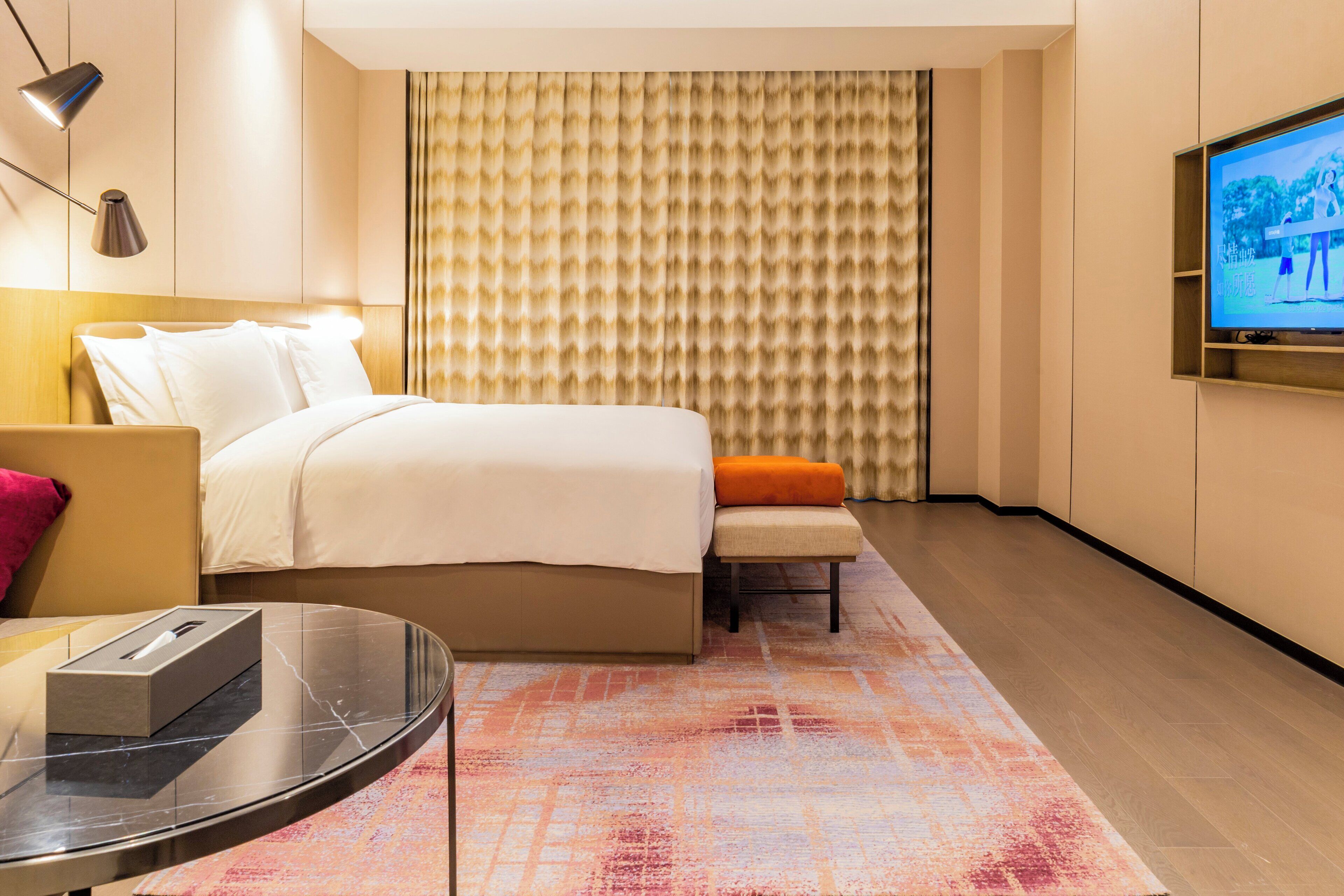 A modern hotel room with a bed, a small table, a bench, a TV, and a patterned carpet.