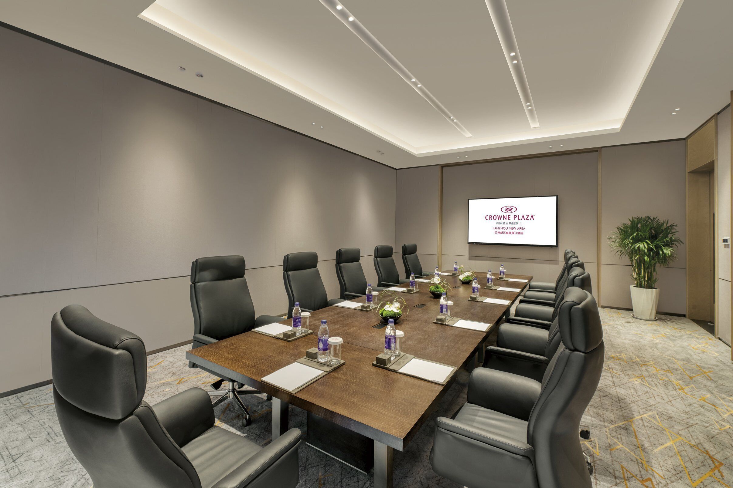A conference room with a long table, black leather chairs, water bottles, and a screen displaying "CROWNE PLAZA".