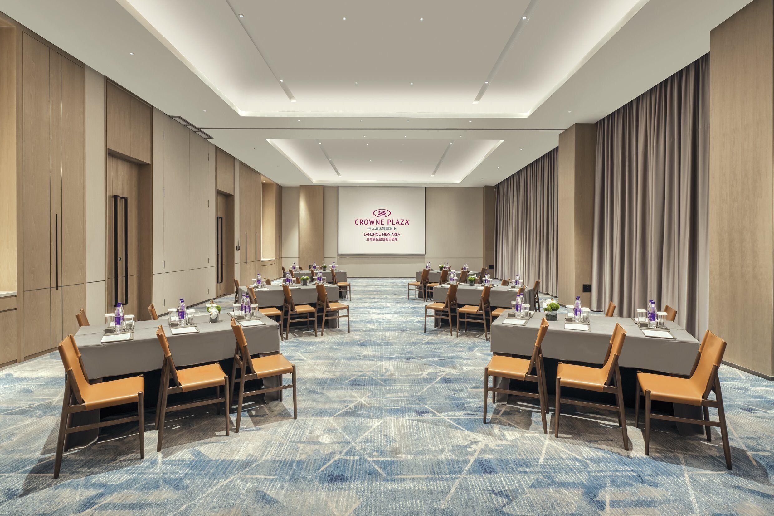 A conference room with a large screen displaying "CROWNE PLAZA" at the front. The room has a long table with chairs, a carpeted floor, and wooden paneling on the walls.