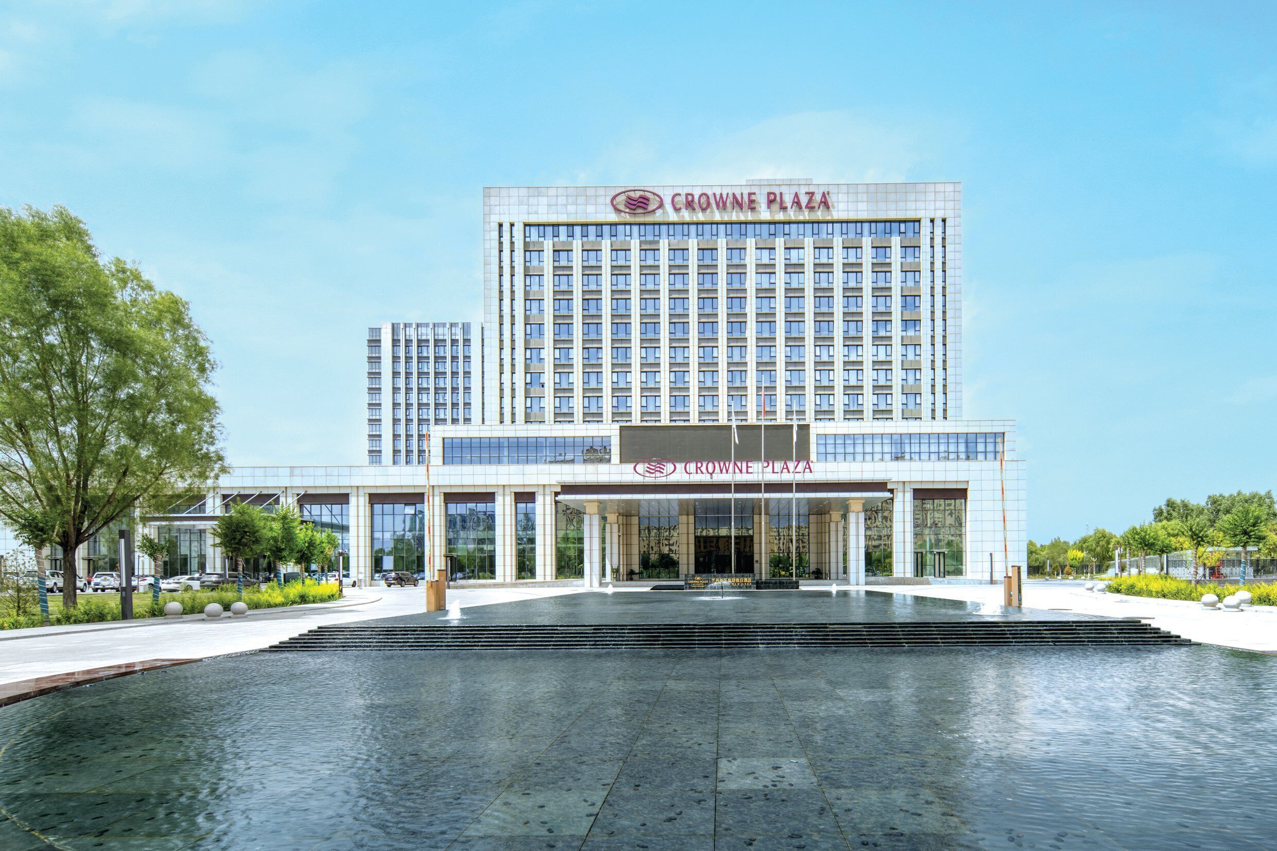 Un hôtel moderne, reconnaissable à son grand panneau « Crowne Plaza », doté d’un hall d’entrée spacieux et agrémenté d’un élément décoratif aquatique réfléchissant à l’avant.