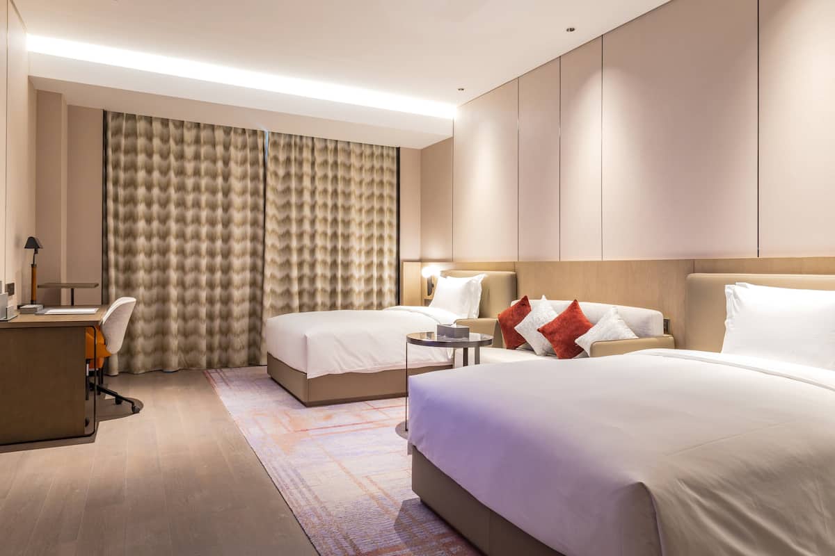Une chambre d’hôtel avec deux lits, un bureau, une chaise, une petite table et une grande fenêtre avec des rideaux.
