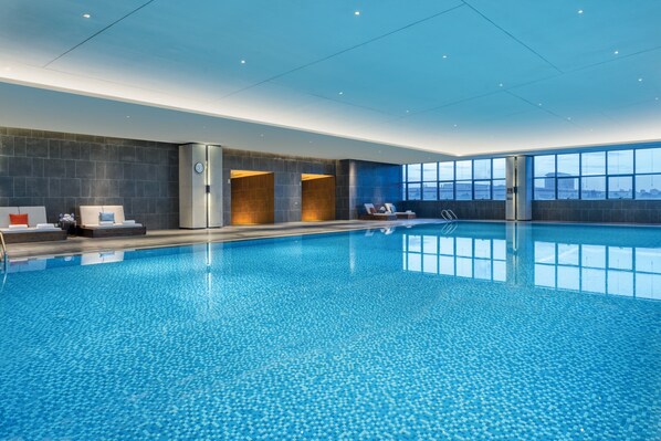 An indoor swimming pool with a tiled floor, a seating area with chairs and a sofa, and a large window offering a view of the city skyline.
