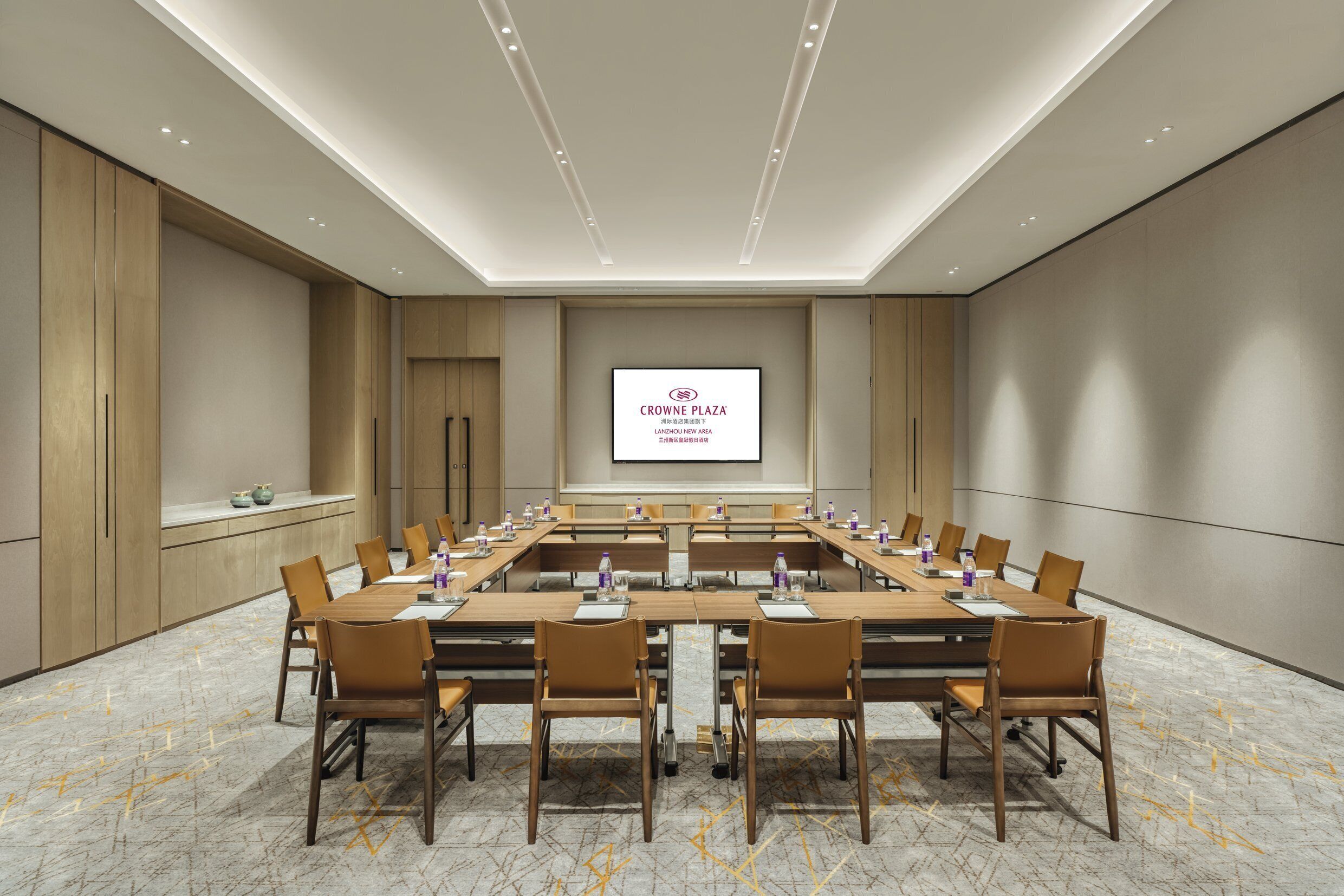 Un salón de conferencias con una larga mesa, sillas, una pantalla de proyector y una presentación sobre Crowne Plaza.