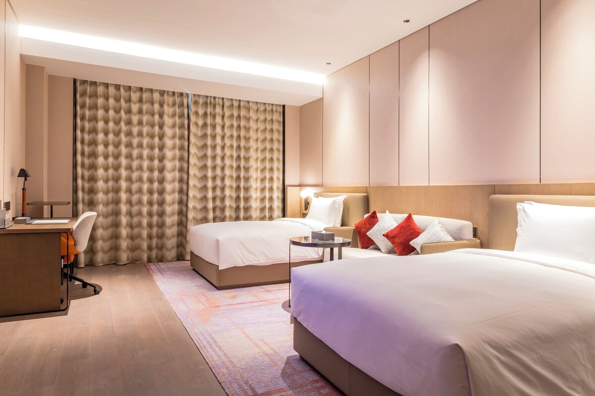 Ein Hotelzimmer mit zwei Betten, einem Schreibtisch, einem Stuhl, einem kleinen Tisch und einem großen Fenster mit Vorhängen.