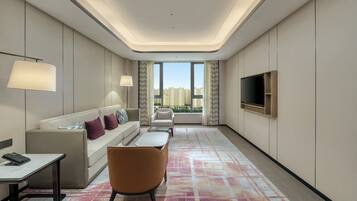 A modern hotel room with a sofa, armchair, TV, and a view of the city through large windows.