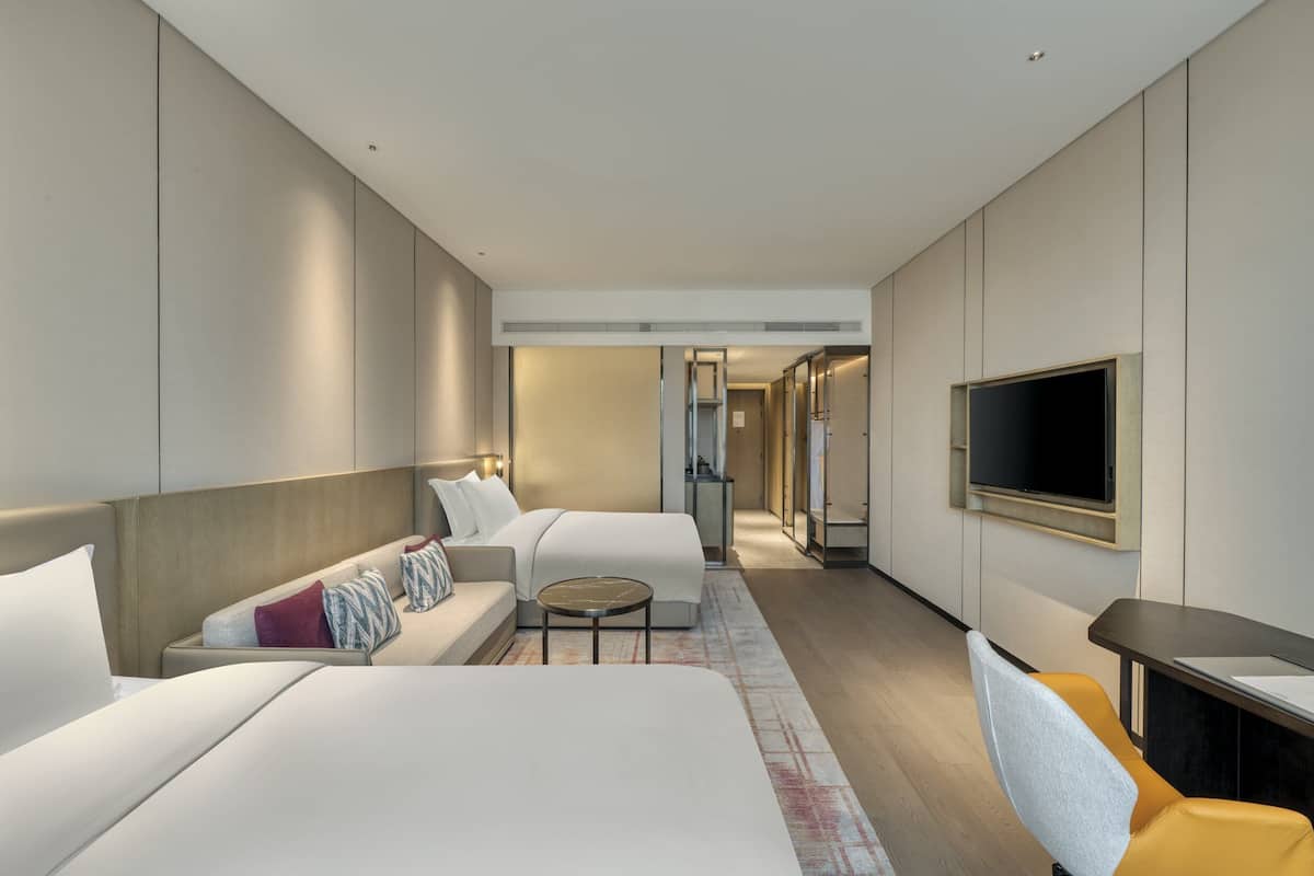 Une chambre d’hôtel moderne équipée de deux lits, d’un canapé, d’une petite table et d’un téléviseur fixé au mur.