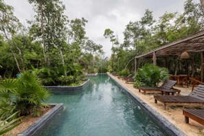 Uma área de piscina com espreguiçadeiras de madeira e um espaço de estar coberto, tudo cercado por vegetação exuberante.