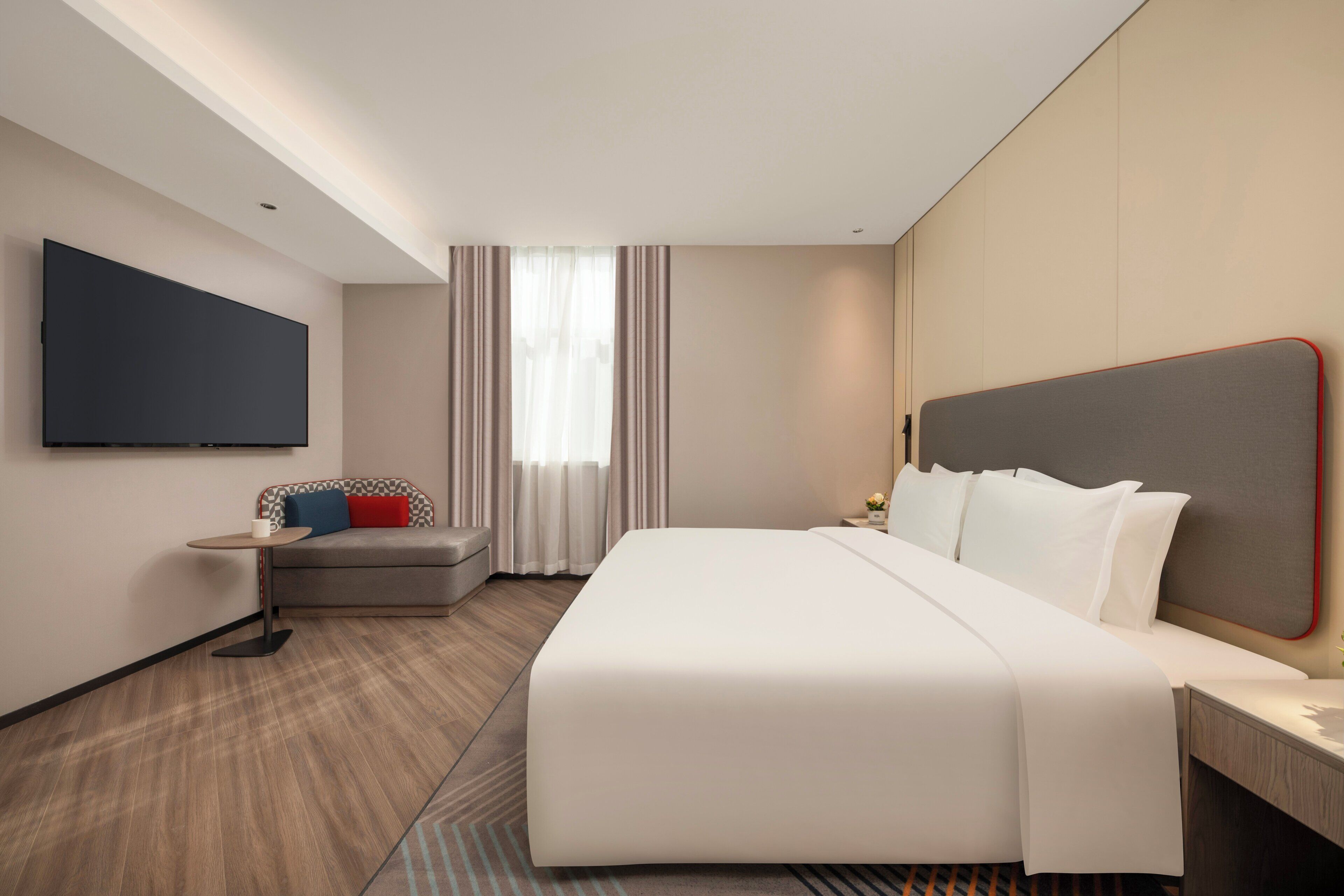 A hotel room with a large bed, a flat-screen TV mounted on the wall, a small table with a cup, and a window with curtains.