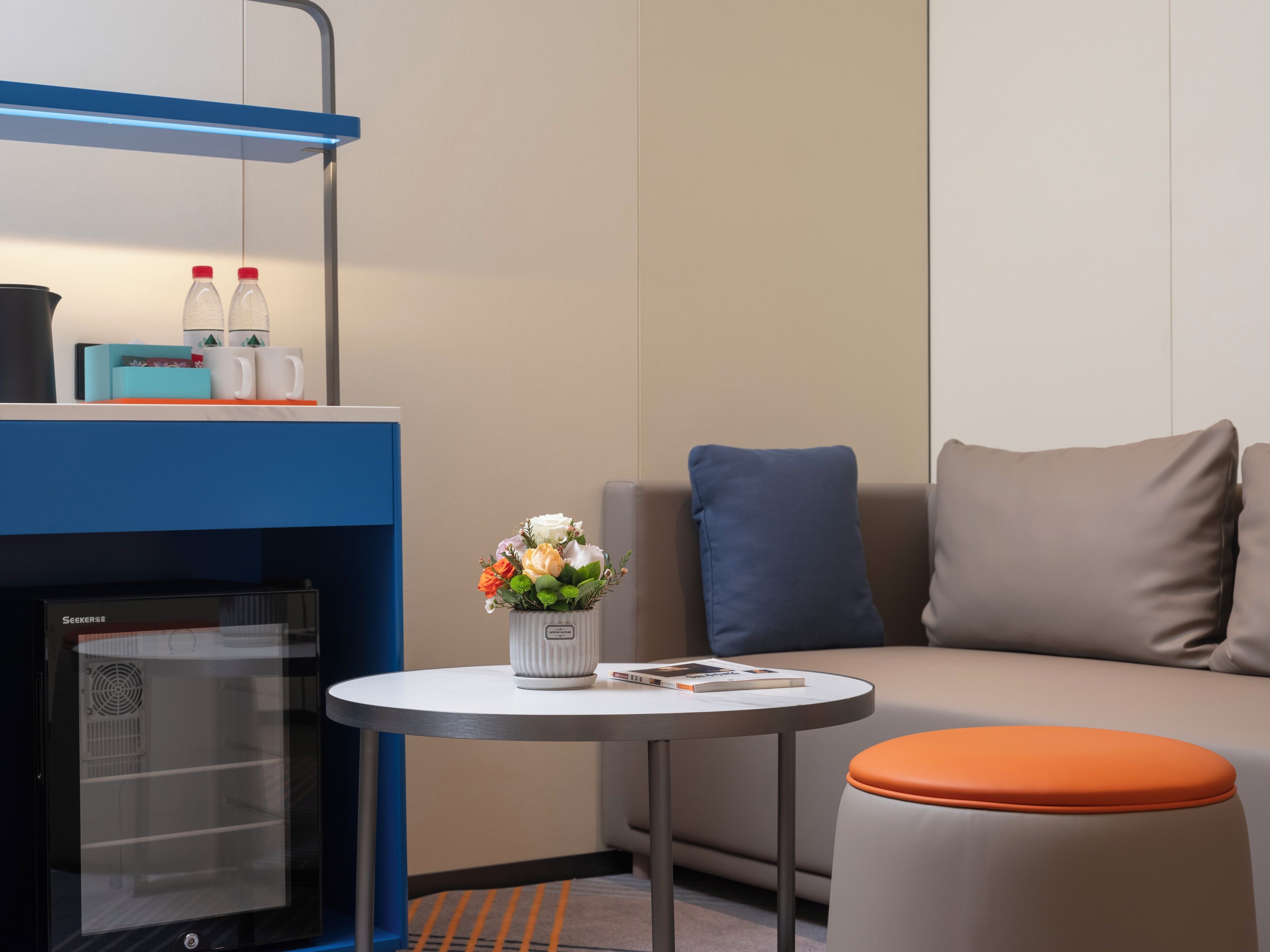 Quarto de hotel moderno com uma escrivaninha azul, uma mesa redonda com um vaso de flores, um sofá com almofadas laranja e azul, e um minibar.