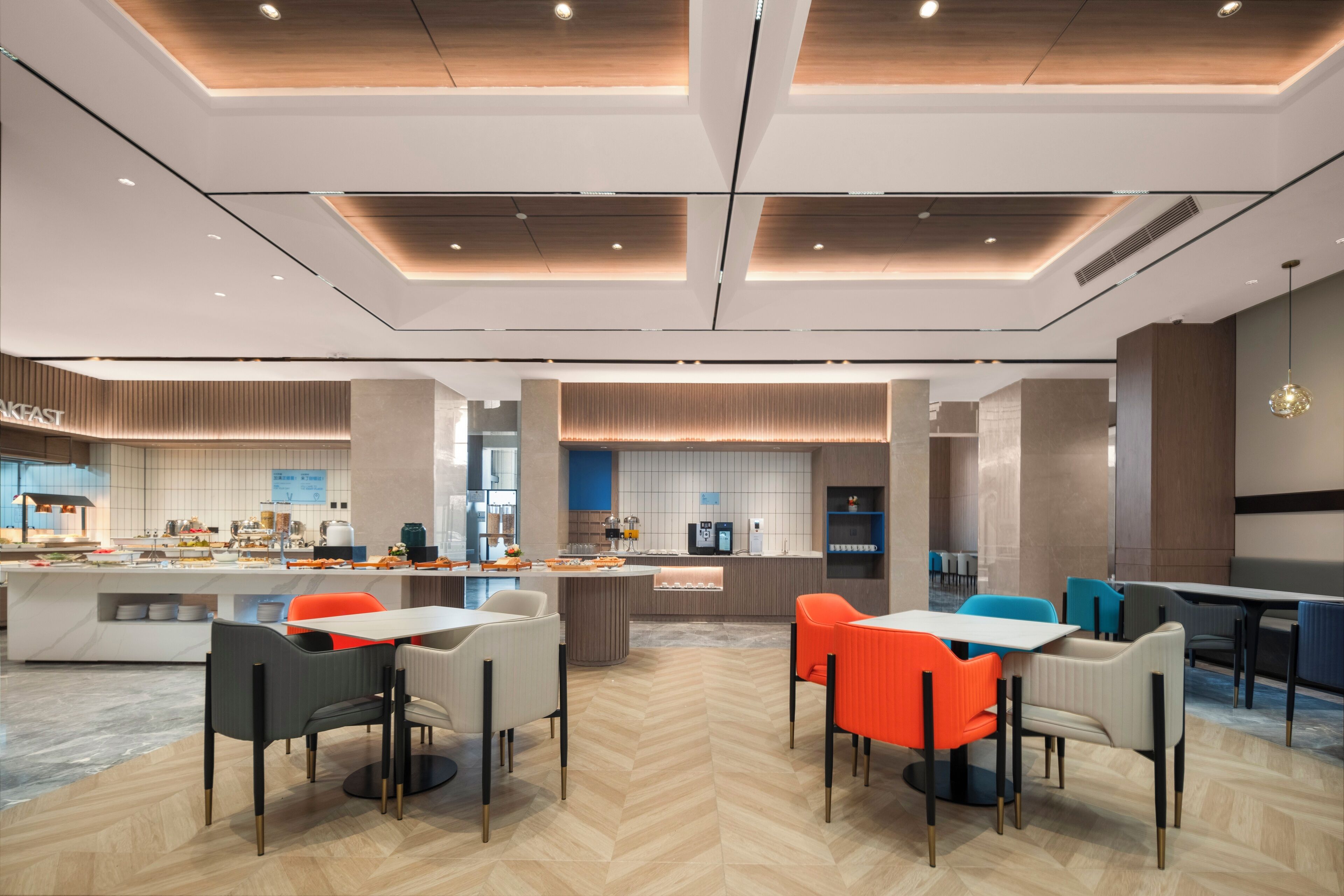 A modern restaurant with a central island, seating areas with orange and grey chairs, and a herringbone floor pattern.