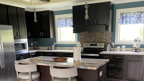 A modern kitchen with dark wood cabinets, a central island with stools, stainless steel appliances, and a window with patterned curtains. - Terrific 4-bedroom house in Tylertown with WiFi, AC (Tylertown)