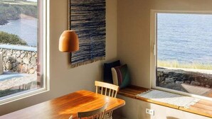 A dining area with a wooden table and chairs, a hanging pendant light, and a view of the sea through large windows.
