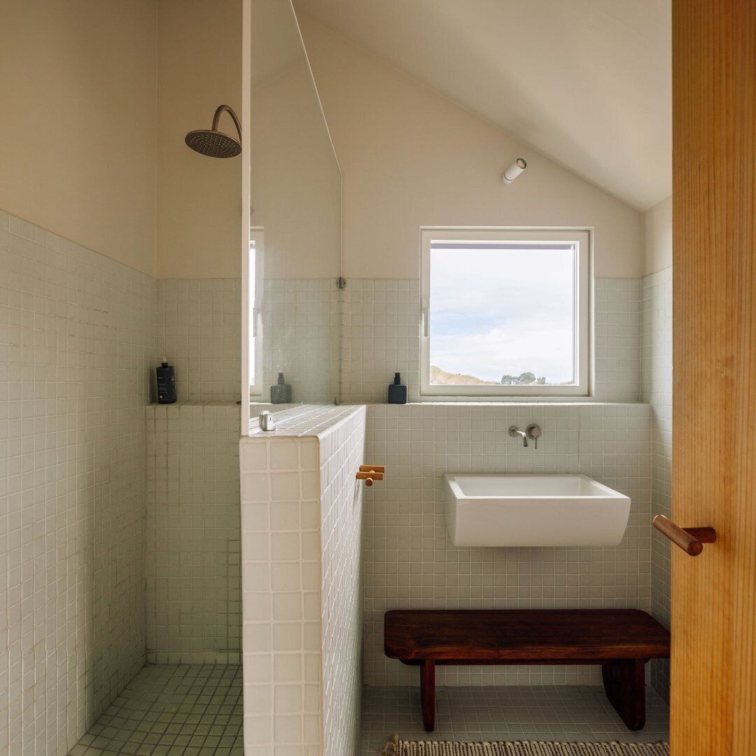 A bathroom with a shower, a sink, a window, and a bench.