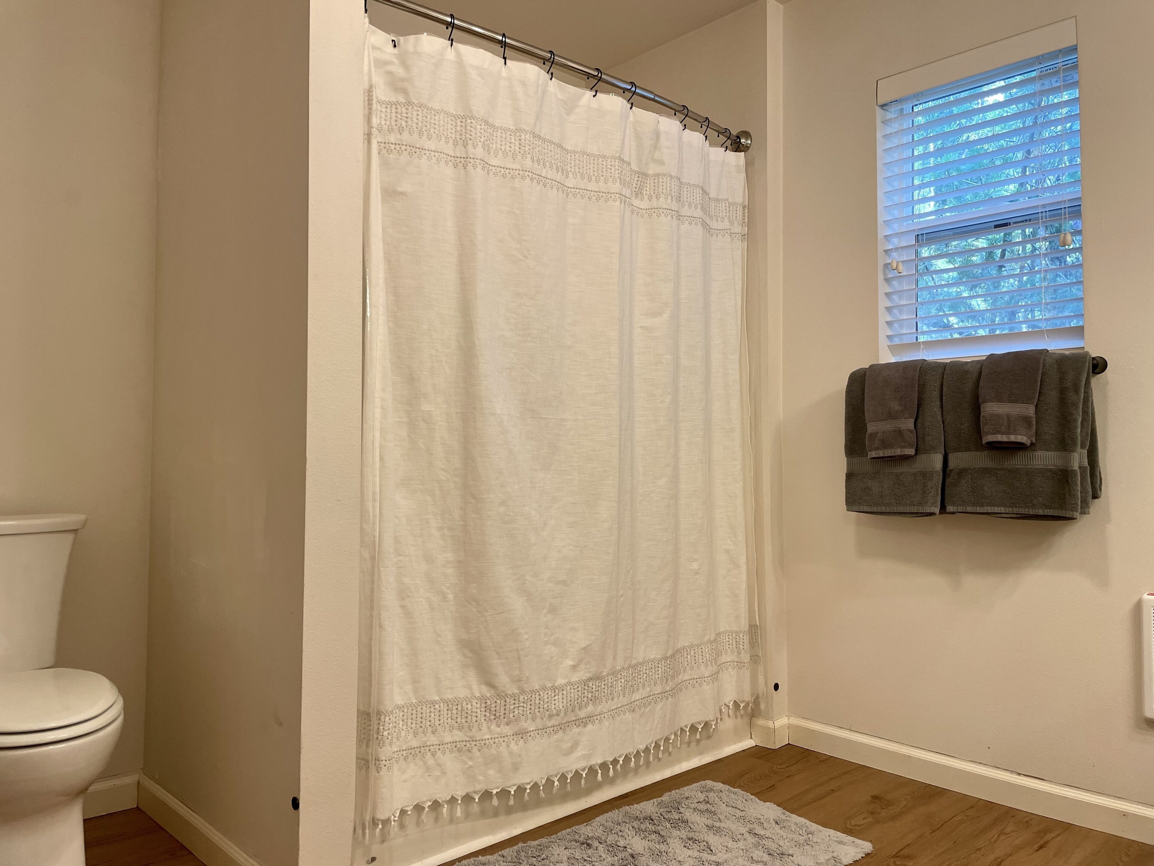 Un baño con cortina de ducha blanca, un inodoro, una ventana con persianas y dos toallas colgadas en la pared.