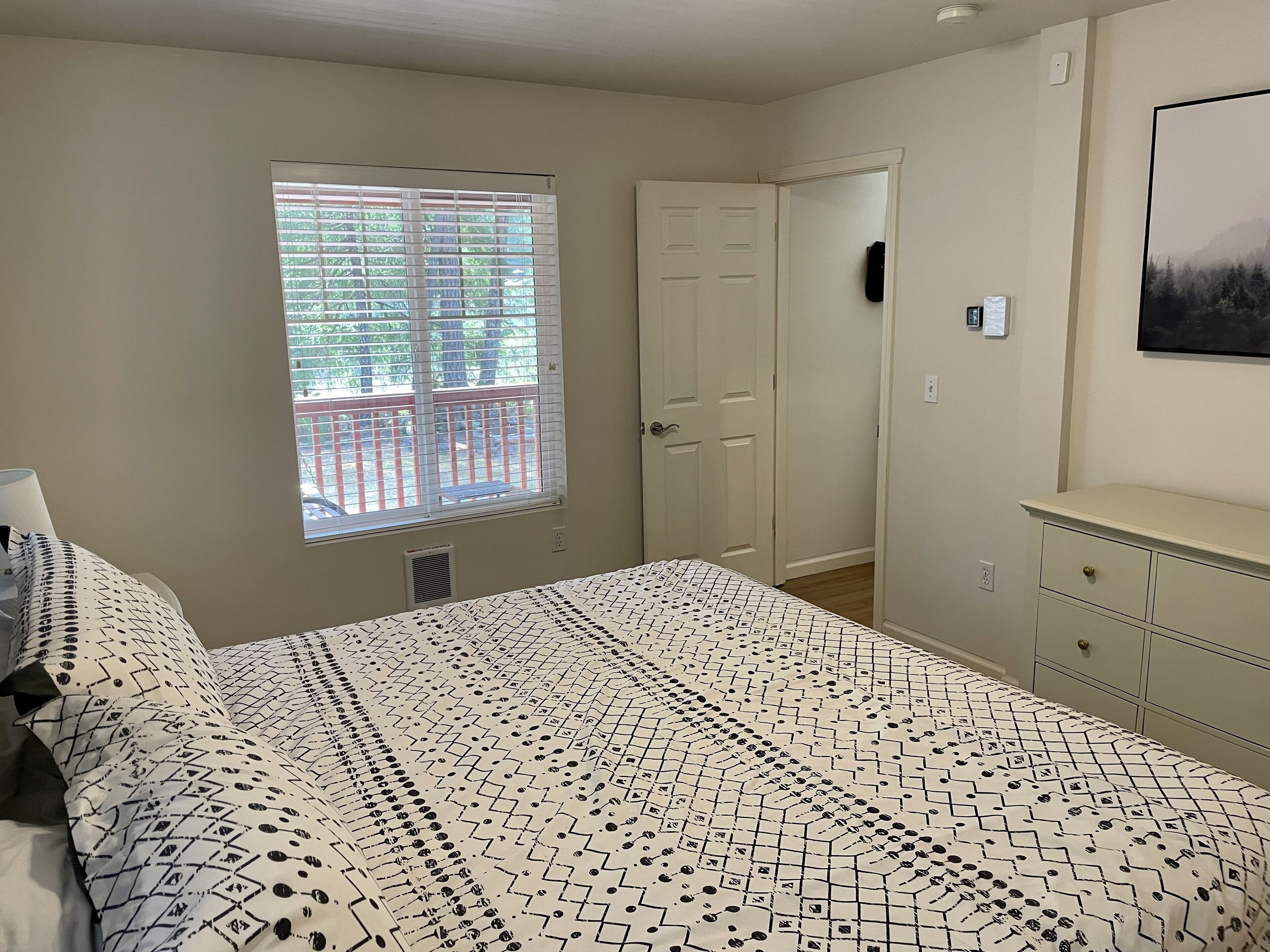 Une chambre à coucher avec un lit, une fenêtre avec des stores, une porte et une commode.
