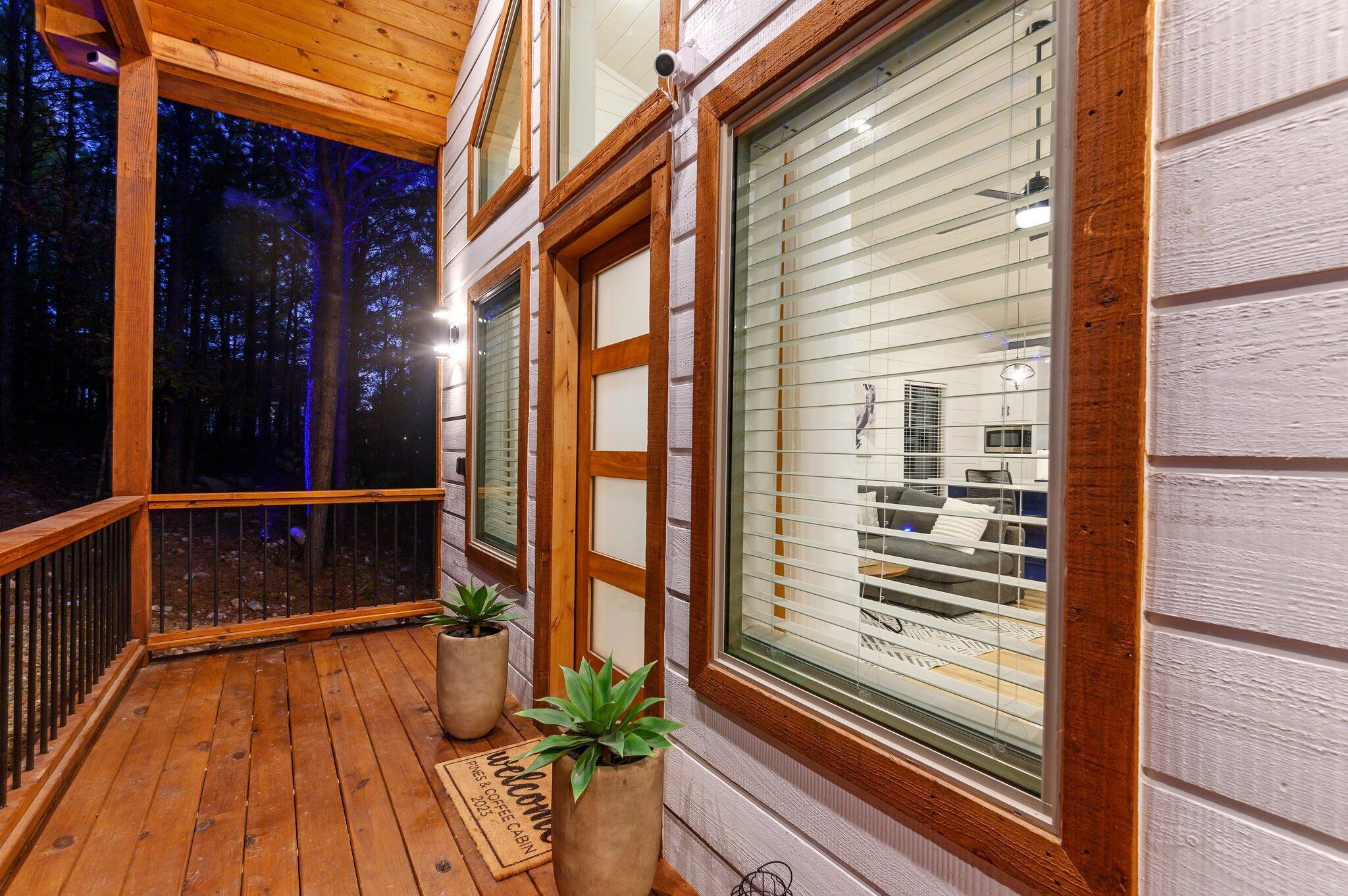 A wooden deck with potted plants, a doormat, and a view of a building through large windows.