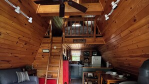 Interior acogedor de una cabaña con sofá, alfombra estampada, mesa de comedor de madera, área de cocina y chimenea.