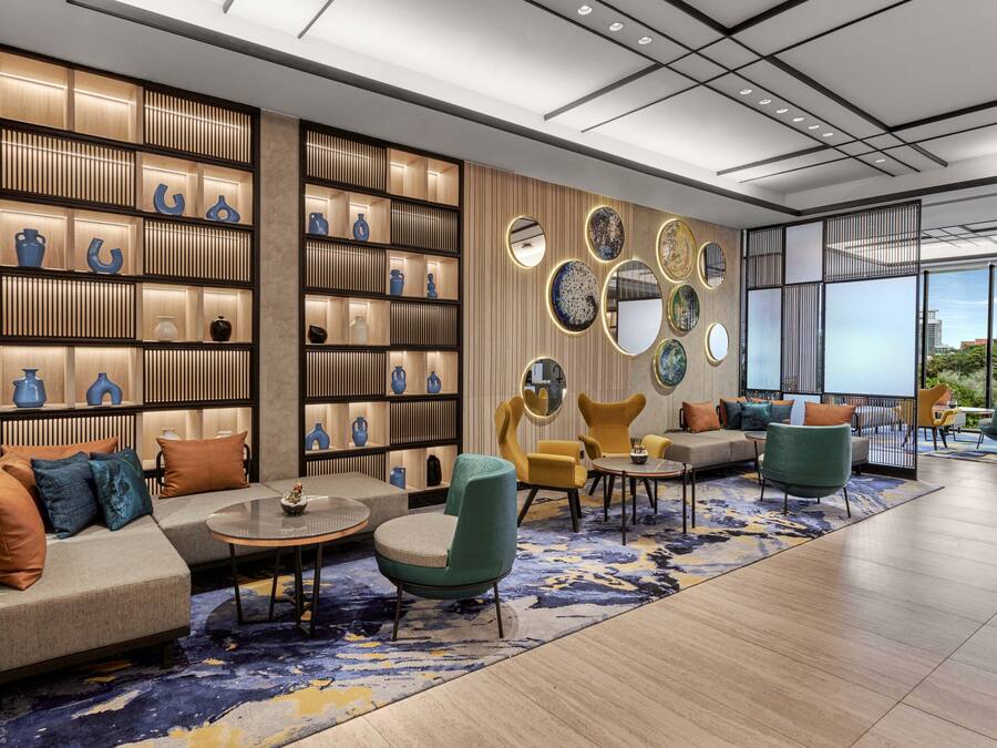 A modern lounge area with a sofa, armchairs, and a coffee table. There are shelves with decorative items and a large window with a view.
