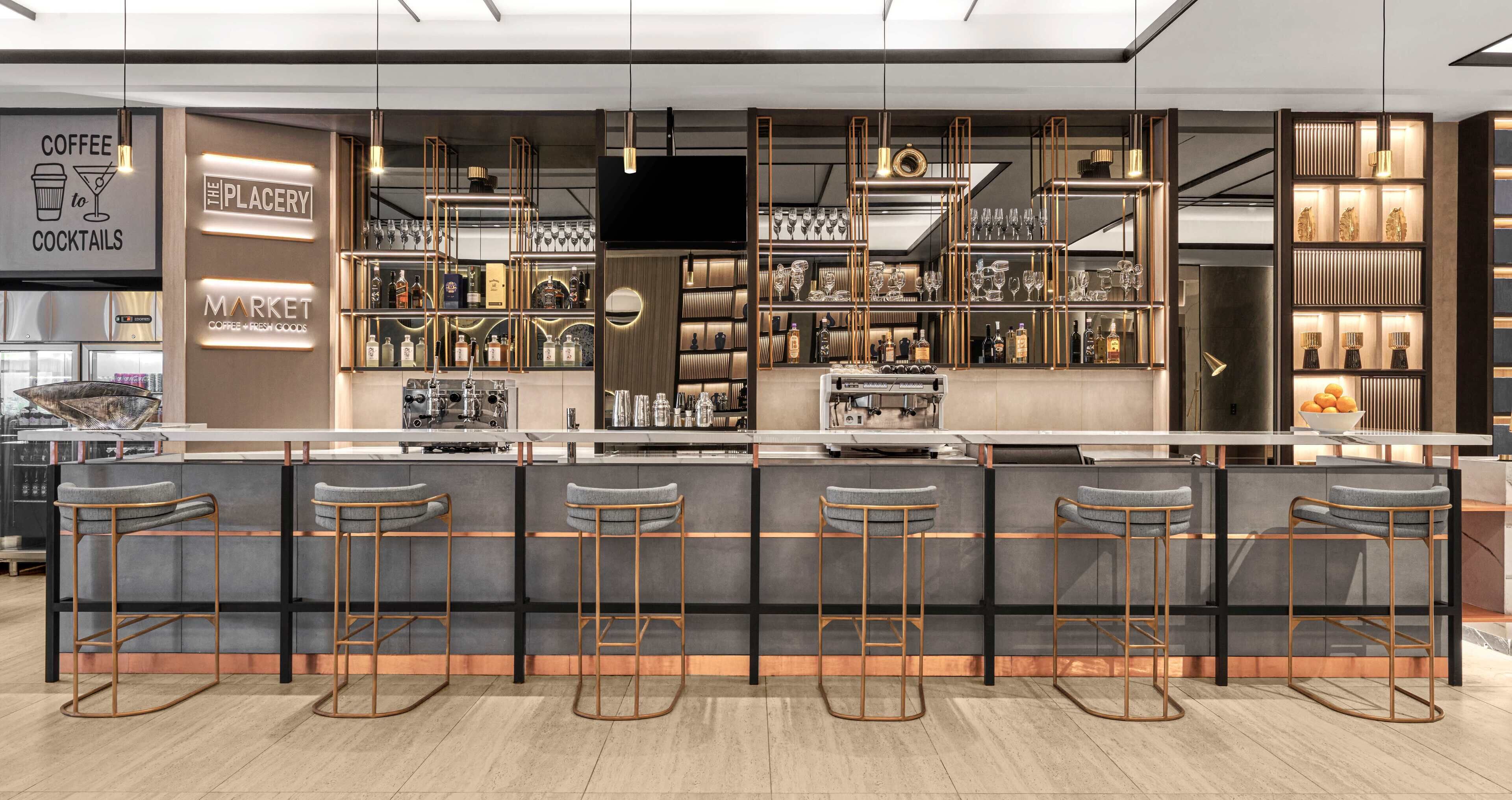 Un área de bar moderna con una barra alargada, varios taburetes y estantes llenos de copas y botellas.