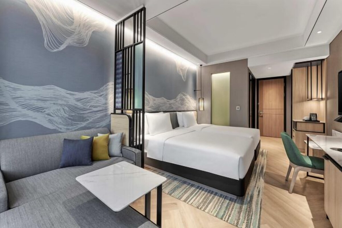 A modern hotel room with a large bed, a sofa, a desk, and a wall with abstract art.