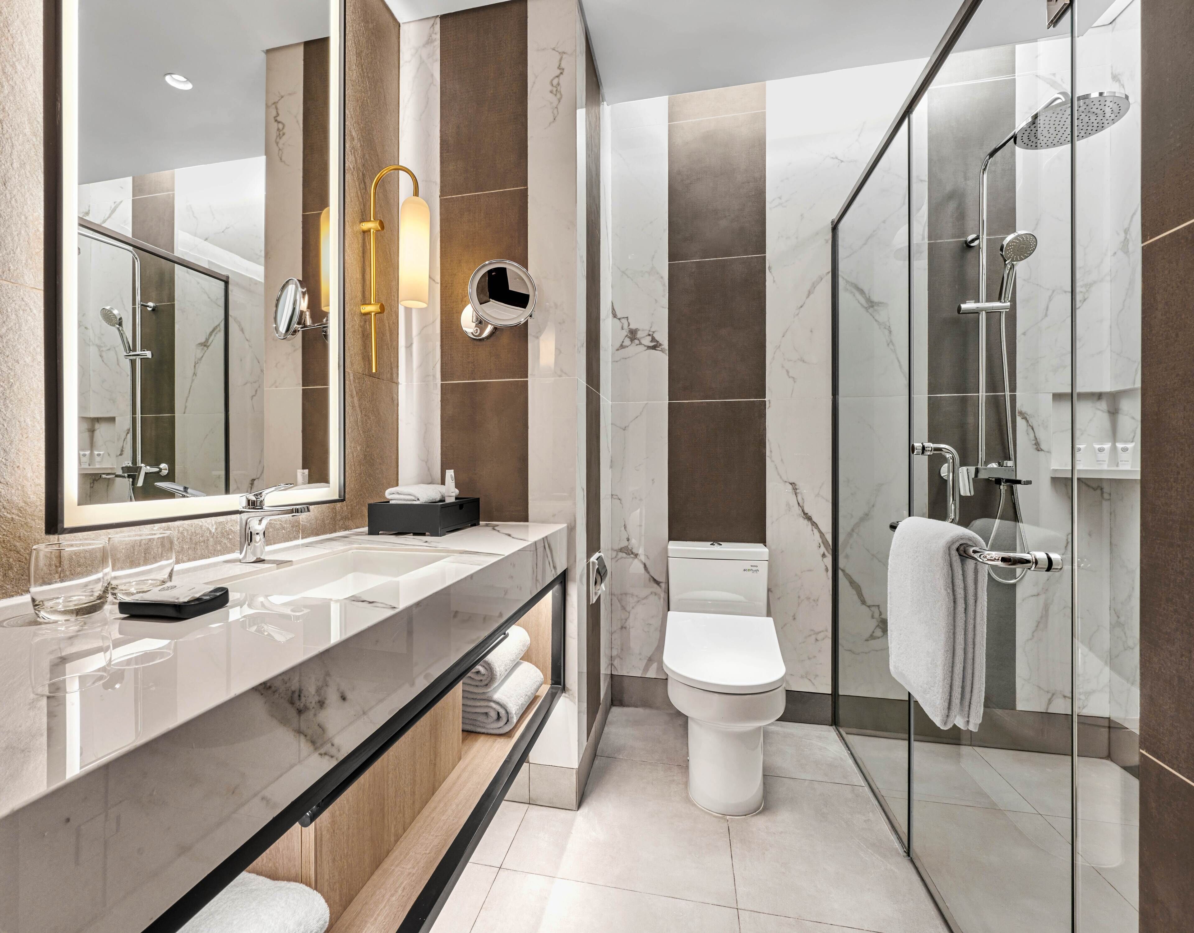 A modern bathroom with a large mirror, a glass shower enclosure, a toilet, and a vanity with a sink and towel rack.
