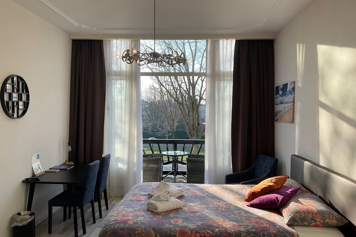 A hotel room with a bed, a desk, a chair, a balcony, and a view of trees.