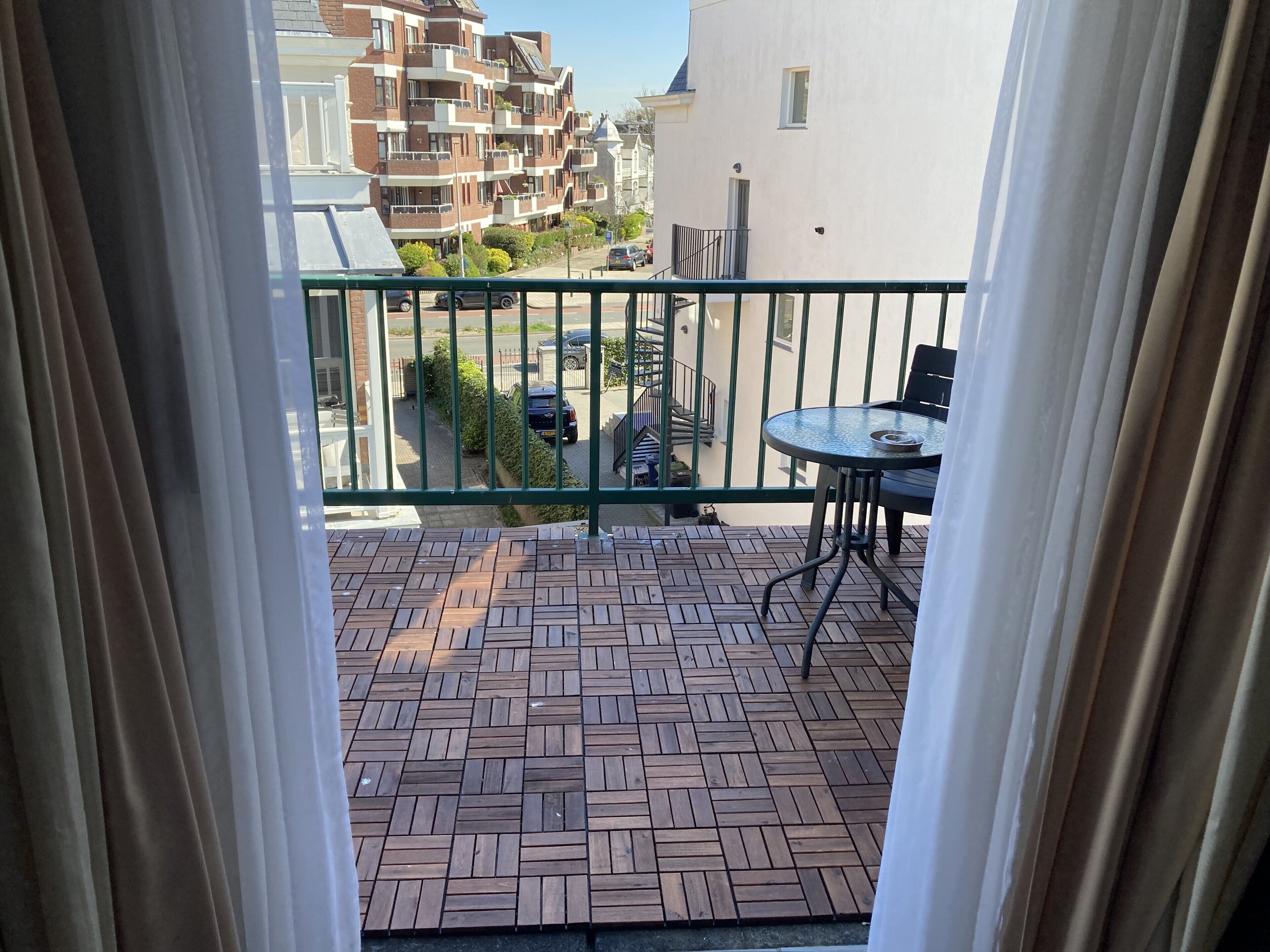 A balcony with a patterned floor, a table, and chairs, overlooking a street with cars and buildings.