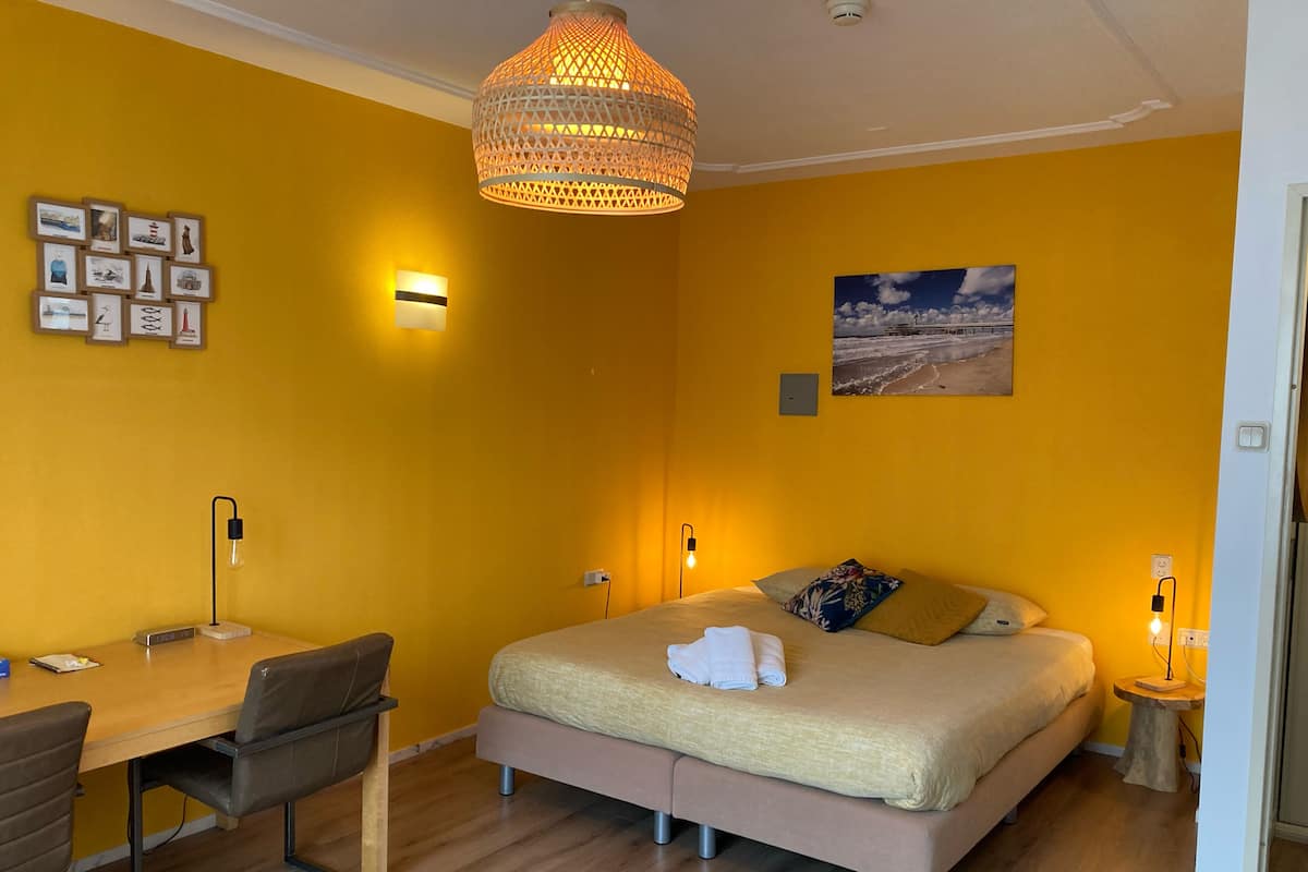 A room with a bed, bedside table, lamp, and a desk with a chair.