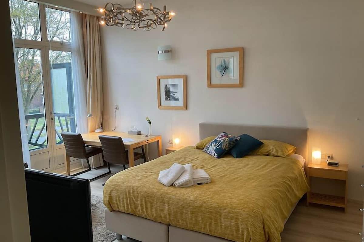 A bedroom with a bed, bedside tables, a desk, and chairs. There are two framed pictures on the wall, and a chandelier hangs from the ceiling.