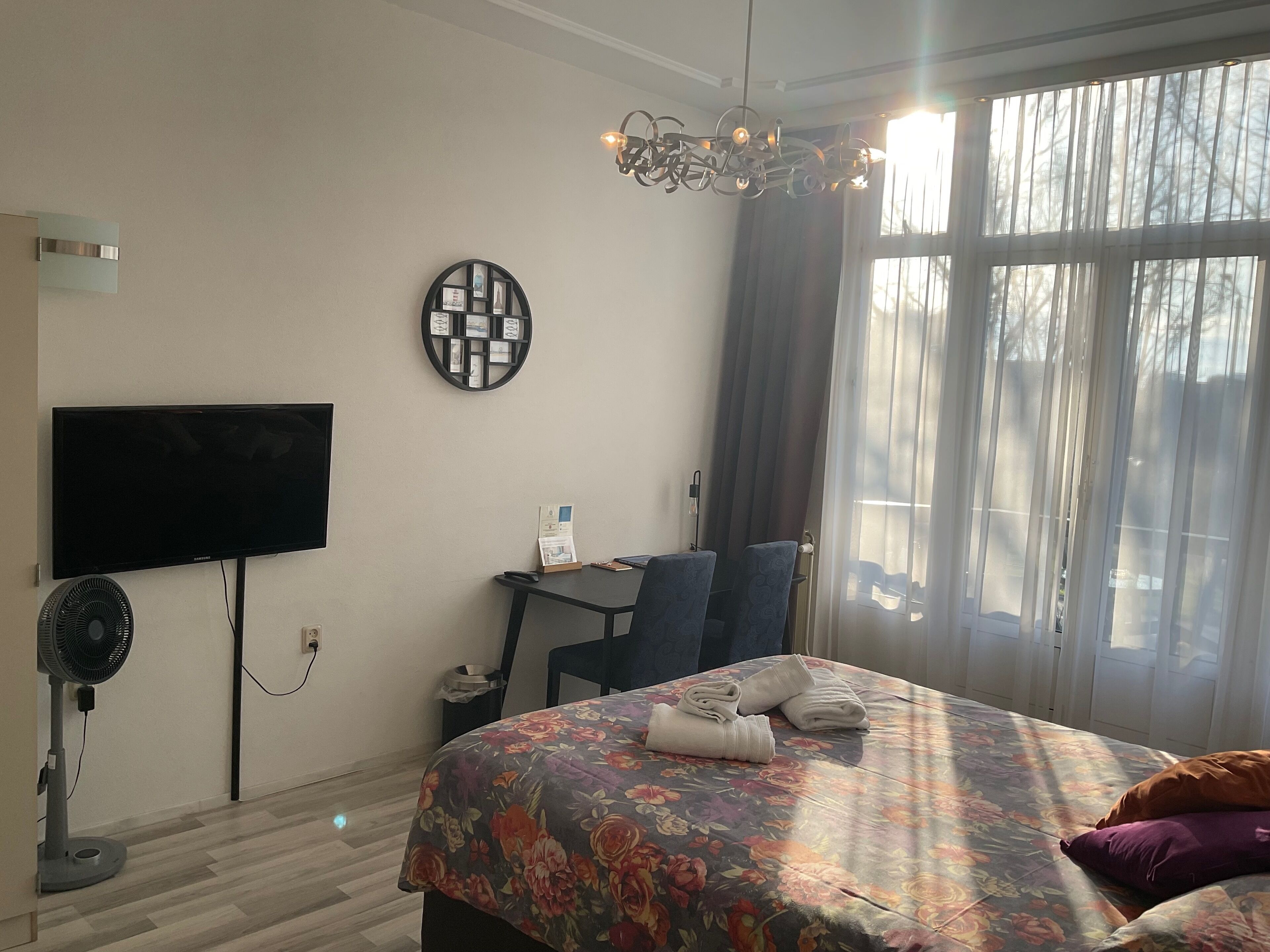 A hotel room with a bed, a desk, a chair, a TV, a fan, and a window with curtains.