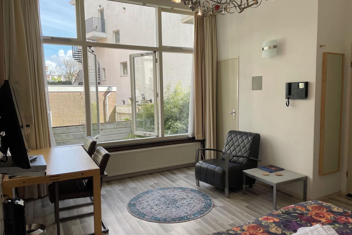 Ein modernes Zimmer mit einem großen Fenster, einem schwarzen Ledersessel, einem kleinen Tisch, einem Bett mit blumigem Überwurf und einer Heizung.