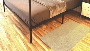 Ein Bett mit Himmel, dessen Gestell aus Holz besteht, eine braune Matratze und zwei Kissen. Ein Nachttisch mit Lampe und Schublade steht daneben. Der Raum verfügt über Holzböden und ein Fenster mit Vorhängen.