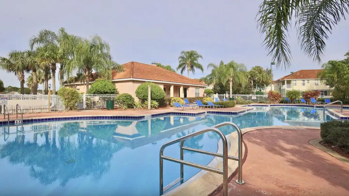Une piscine avec des chaises longues, des palmiers et un bâtiment doté d’un toit en tuiles.