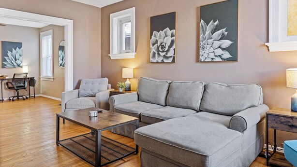 Ein modernes Wohnzimmer mit einem grauen Ecksofa, einem Couchtisch und einem Schreibtisch mit Stuhl. An den Wänden sind zwei Wandleuchten und gerahmte Kunstwerke angebracht.