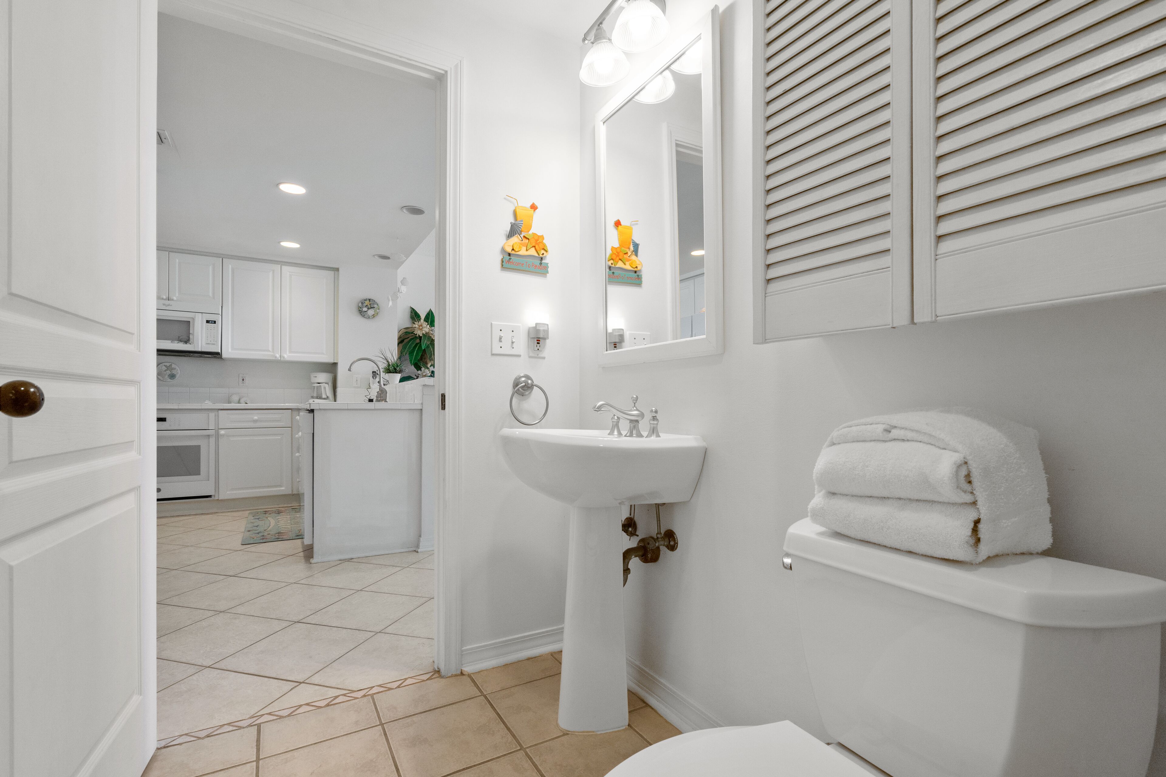 Un baño moderno con un lavabo, inodoro y tocador blancos. Hay un espejo sobre el lavabo y una toalla en el inodoro. El baño está conectado a una cocina con armarios blancos y un microondas.