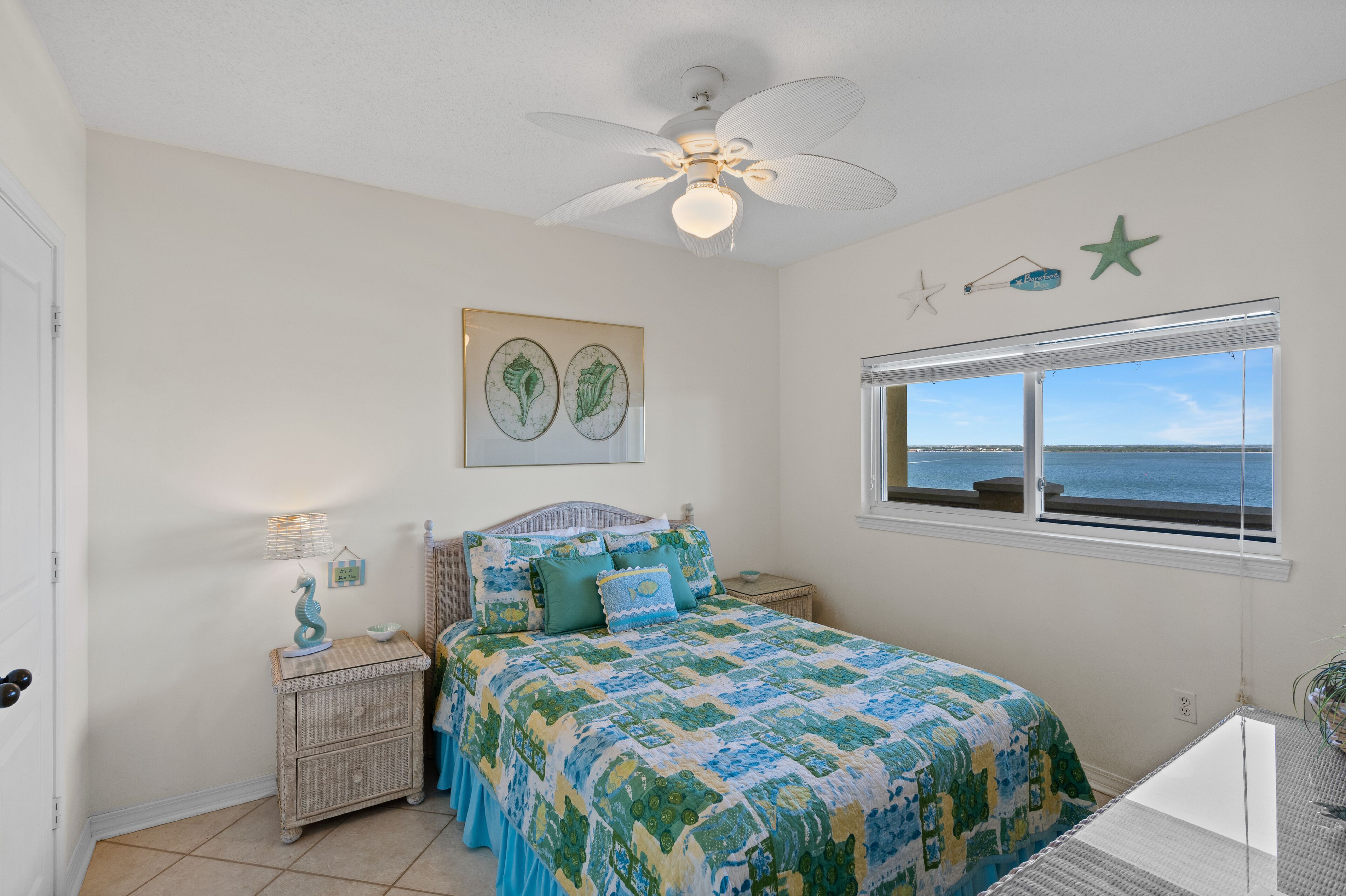Une chambre à coucher comprenant un lit, un ventilateur de plafond, une fenêtre donnant sur la mer et des œuvres d’art décoratives sur les murs.