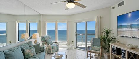 A spacious living room with a large window overlooking the ocean, a ceiling fan, and a bookshelf.