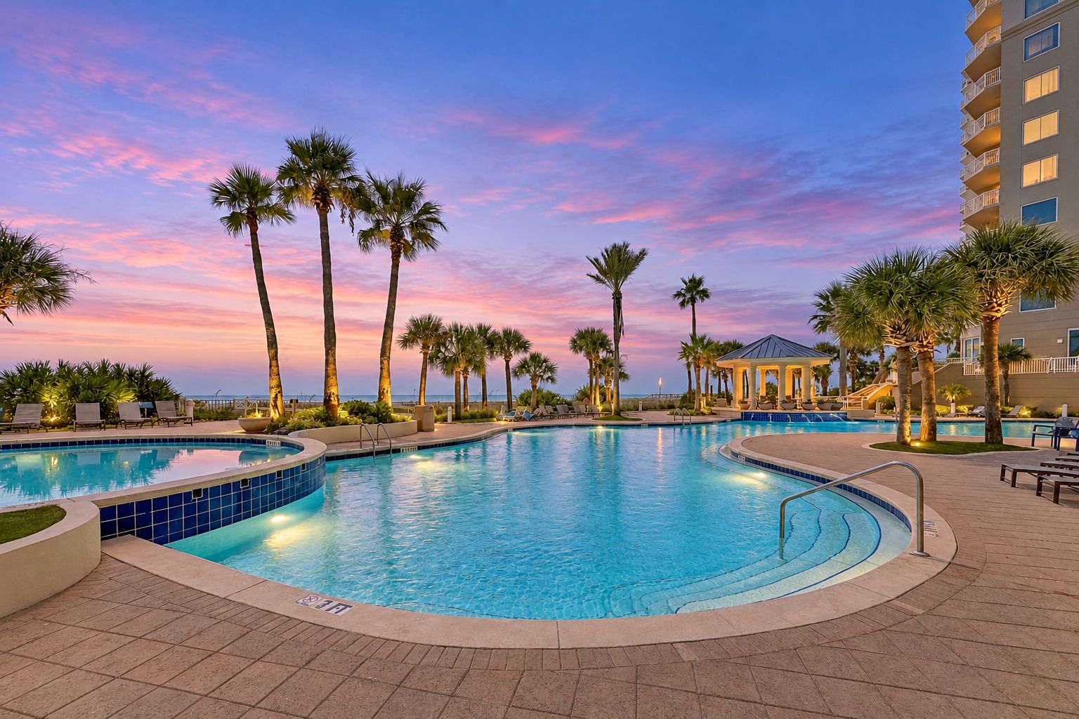A swimming pool with a curved design, surrounded by palm trees and a building with a covered area.