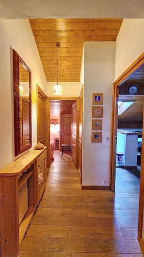 A hallway with wooden flooring and a ceiling with exposed beams. There is a mirror on the left and a wall with framed pictures on the right.