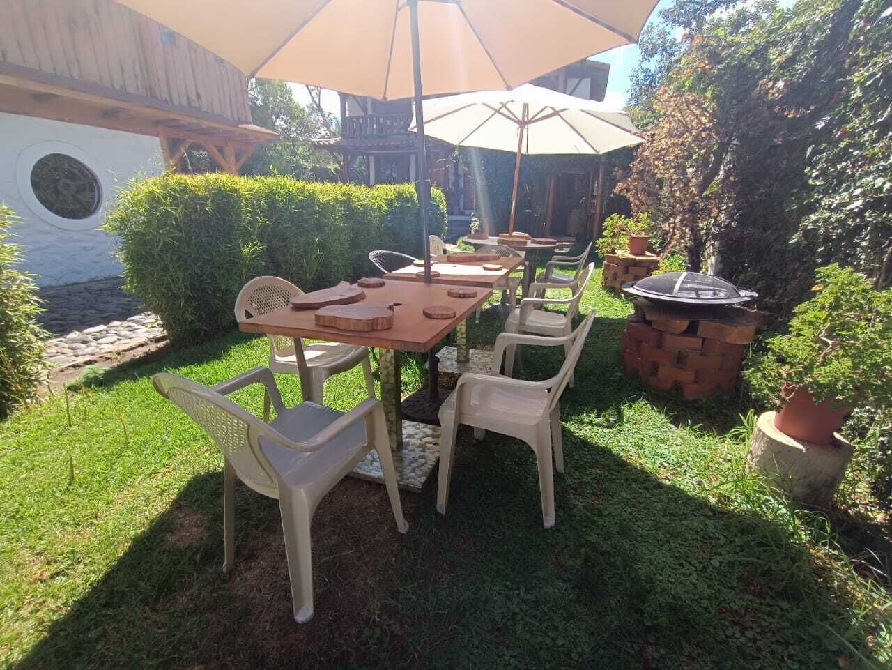Un patio al aire libre con una mesa de madera y sillas de plástico, rodeado de follaje y una estructura de ladrillo.