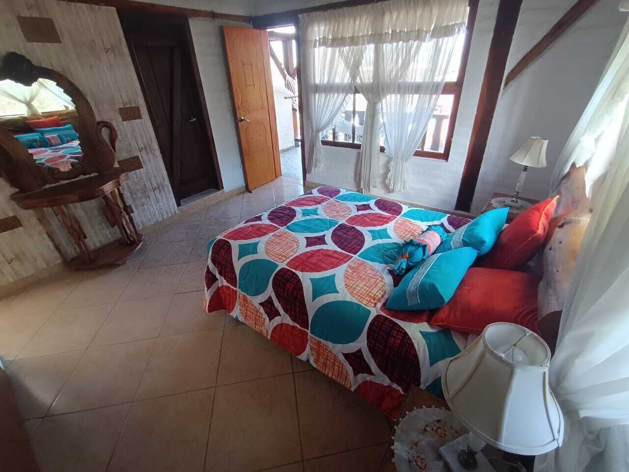 A bedroom with a colorful bedspread, a wooden chair, a lamp, and a view of the outside.