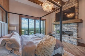 Un dormitorio con chimenea de piedra, un ventanal con vistas y una lámpara moderna.