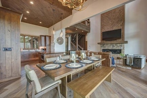 Un comedor espacioso con una larga mesa de madera dispuesta para seis personas, rodeada de asientos cómodos. Se observa una chimenea de piedra y un televisor de pantalla plana grande al fondo.