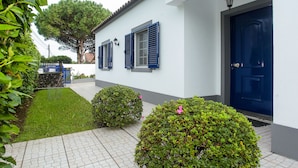 Edifici blanc amb una porta i persianes blaves, un camí empitnat i arbustos verds.