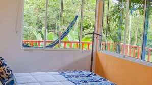 Ein Schlafzimmer mit Bett, Fenster und Blick auf ein rotes Geländer sowie grüne Vegetation.