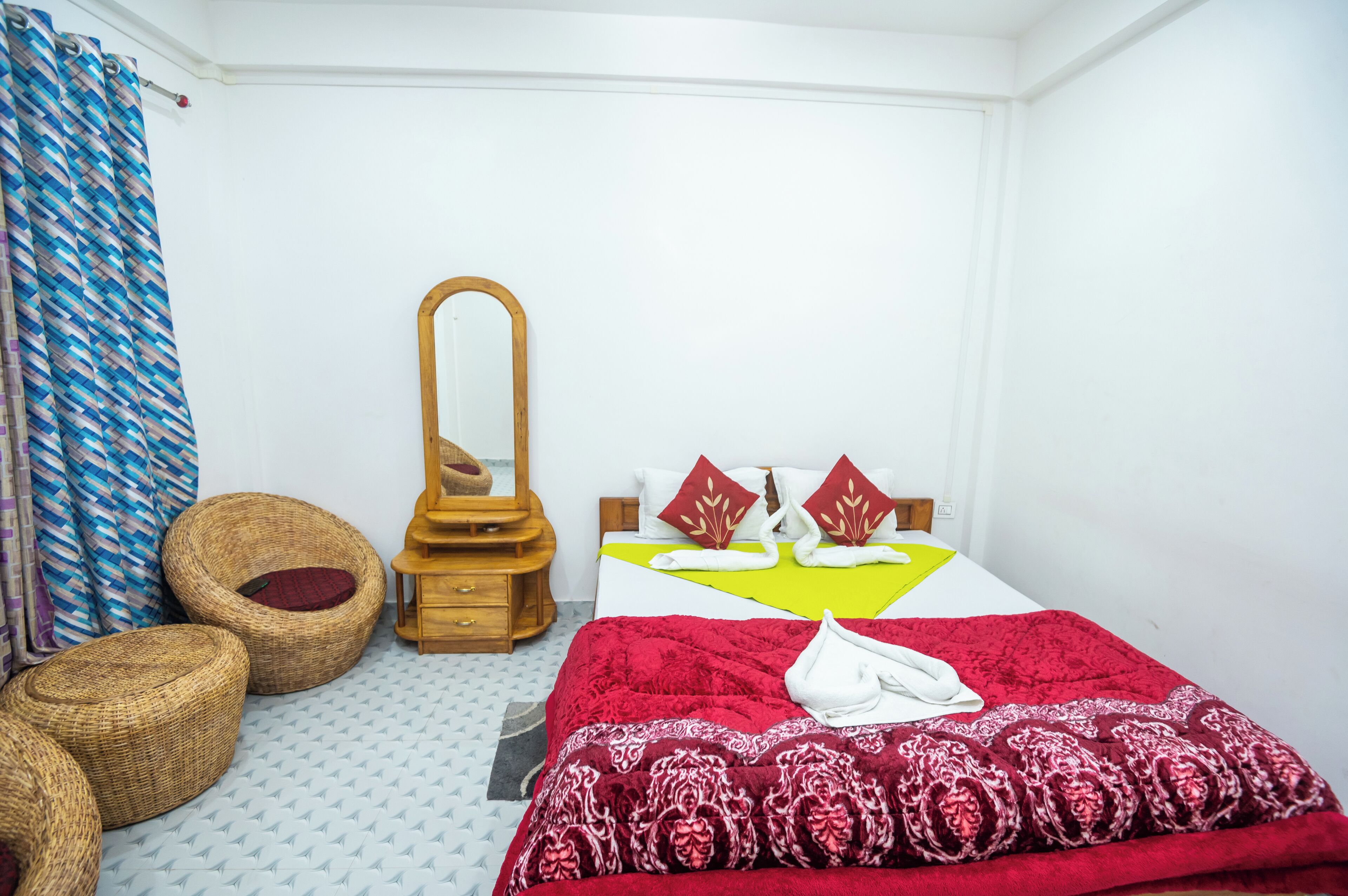 A small, neatly arranged bedroom with a bed, a wooden nightstand, a mirror, and wicker ottomans.