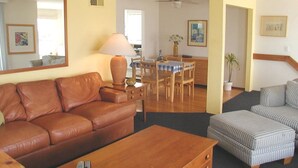 A living room with a brown leather sofa, a wooden coffee table, and striped ottomans. There is a dining area with a table and chairs, a lamp, and a ceiling fan.