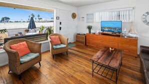 A room with a wooden floor, wicker chairs with cushions, a wooden table, a TV, and a view of the beach through a window. - Cute and cozy one-bedroom unit in South Mission (San Diego)